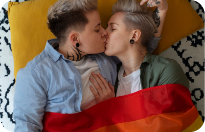 Stock photo of 2 women lying down, wrapped in a rainbow flag, kissing with eyes closed to symbolize a lesbian hookup.
